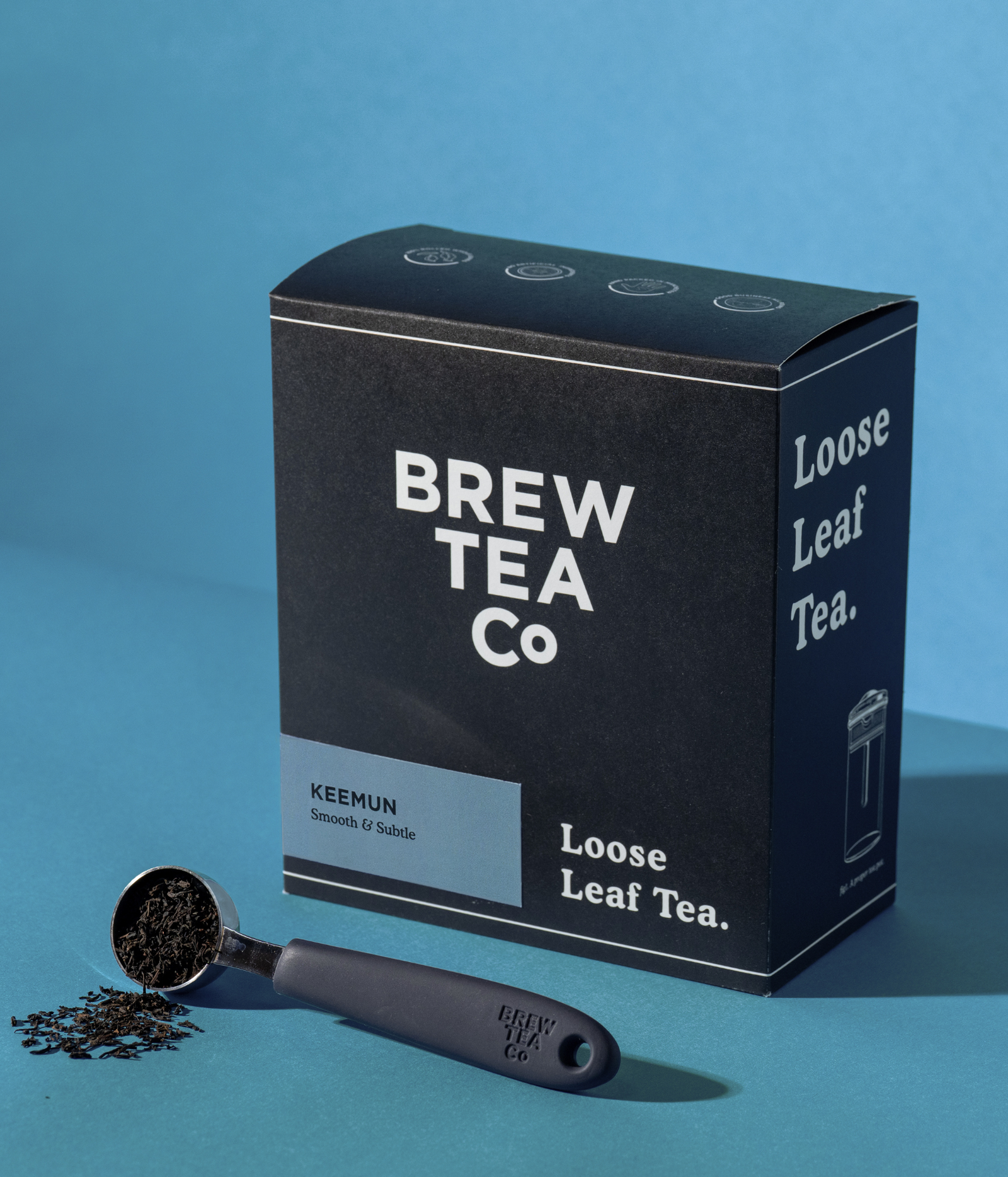 Brew Tea Co. Loose Leaf Tea box of Keemun Tea in black packaging with white text, shown on a blue background with a measuring scoop of loose tea leaves in front.