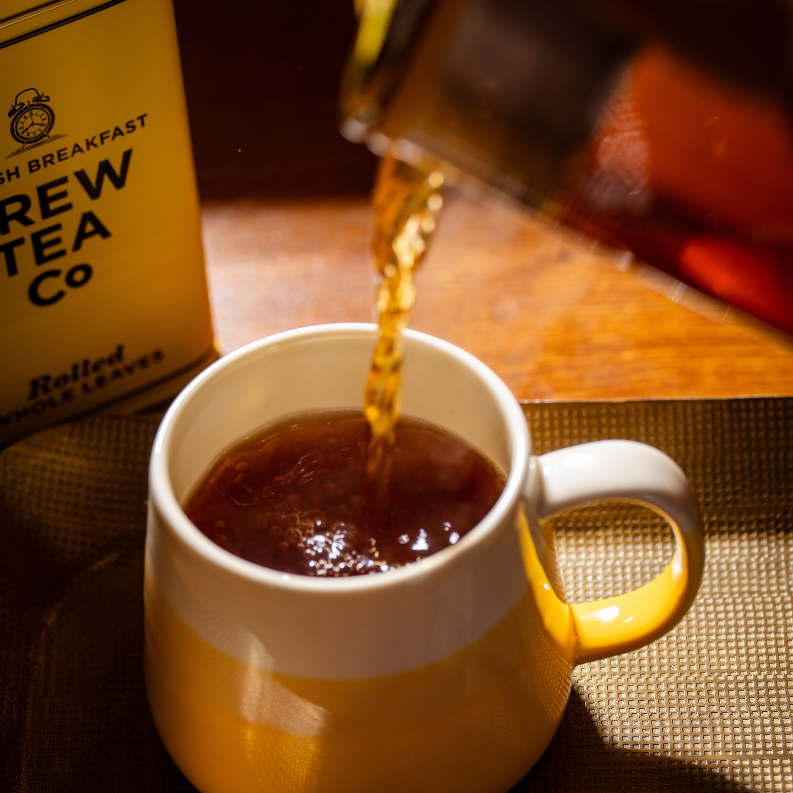 A close-up of brewed tea being poured from a tea pot for one into a white ceramic mug with a yellow lower half. A yellow tin labeled Brew Tea Co is visible in the background on a wooden table.
