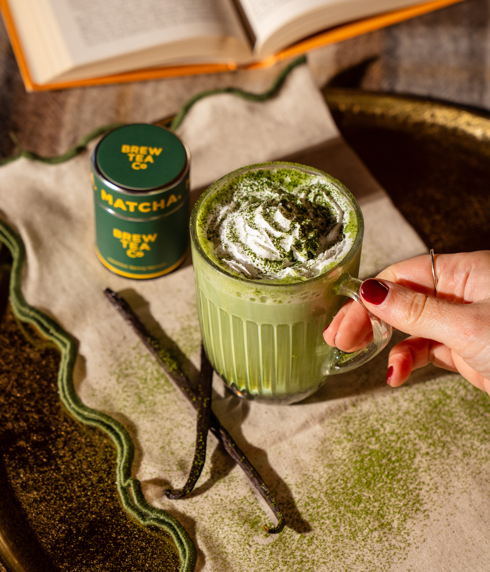 A hand holds a glass of green matcha latte topped with whipped cream and matcha powder, next to a green Brew Tea Co matcha tin, with vanilla beans on a styled tabletop.
