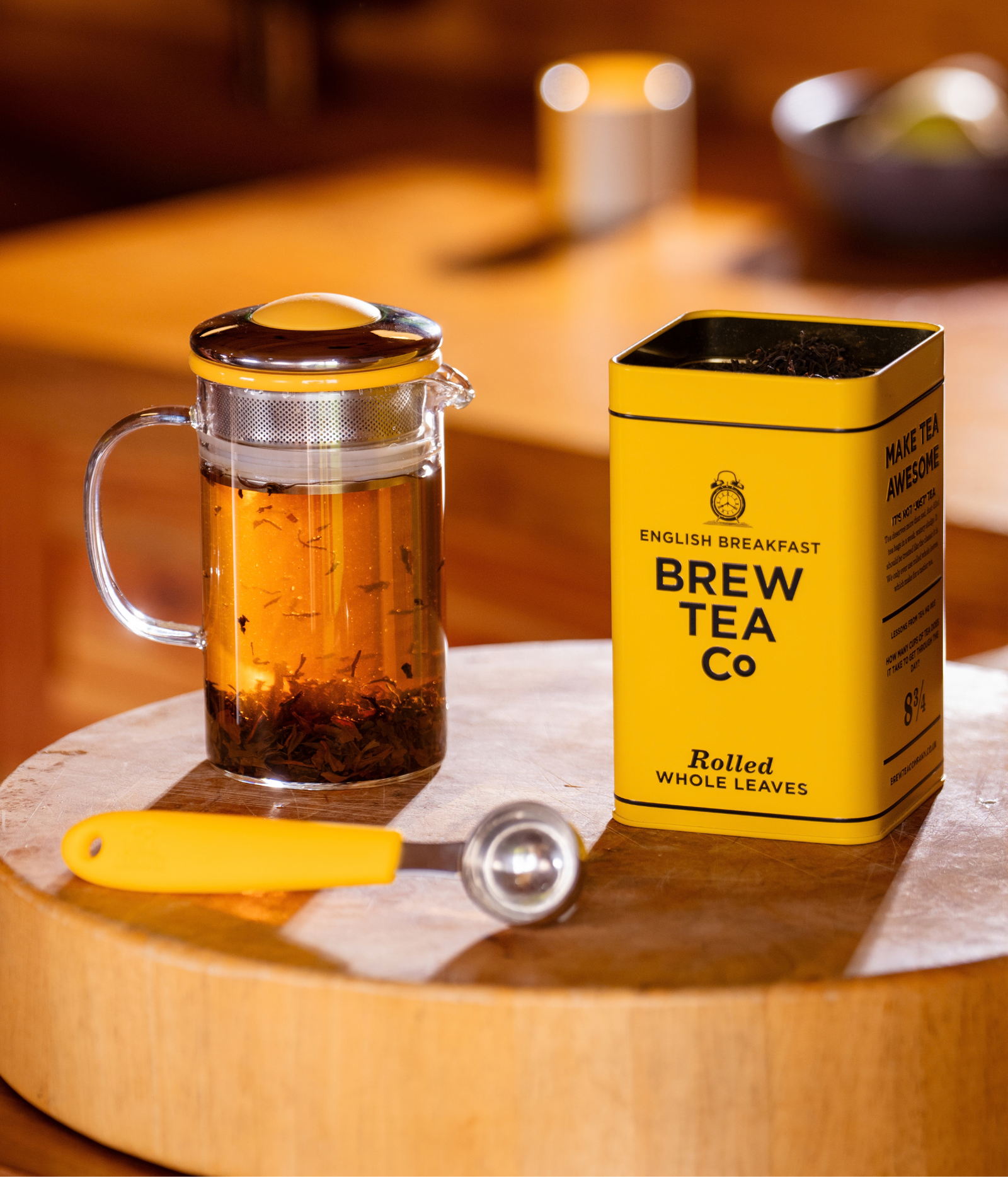 A glass teapot with a yellow lid sits beside a yellow tin labeled 'BREW TEA Co' and 'English Breakfast Rolled Whole Leaves,' on a wooden board with a yellow-handled scoop in a warm indoor setting.